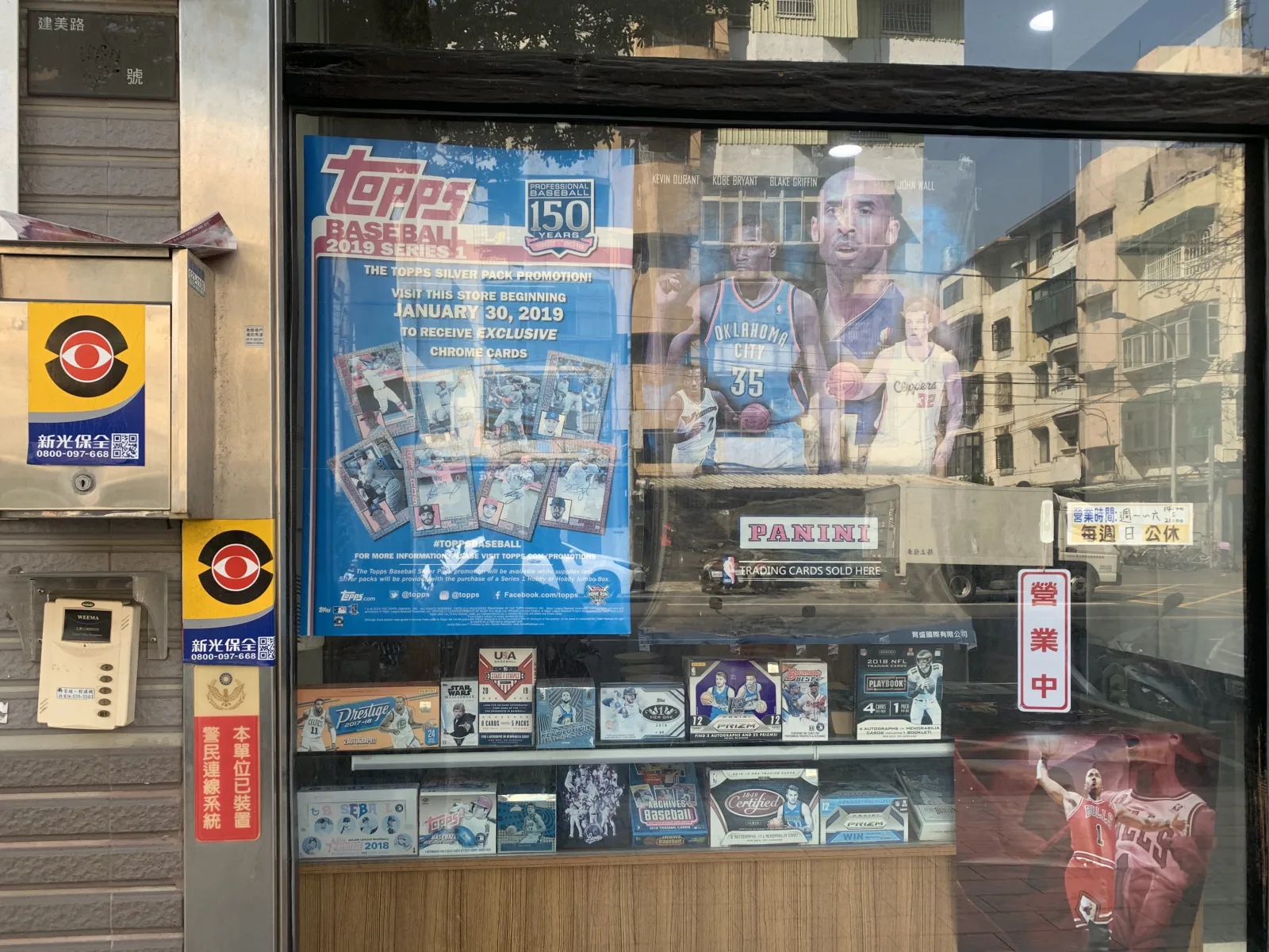 A trading card shop window in Hsinchu City, Taiwan, displaying hobby boxes and card posters.