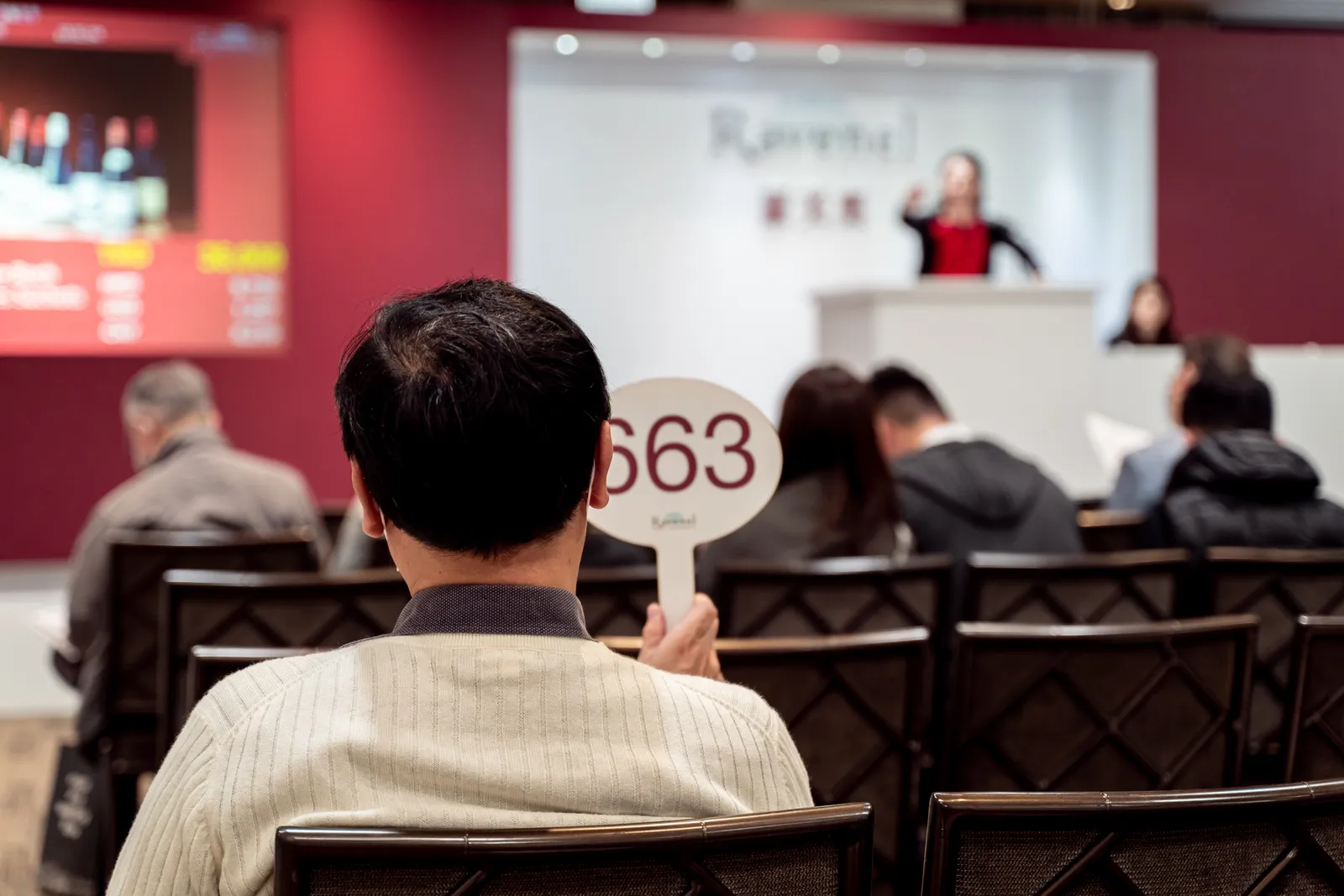 A seated bidder raises a numbered paddle in an auction room while the auctioneer takes bids from a rostrum.
