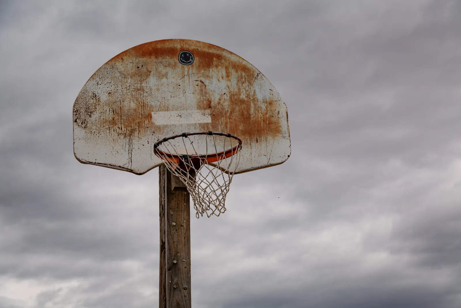 A weathered, rusted basketball hoop and backboard mounted on a wooden pole against a cloudy sky.
