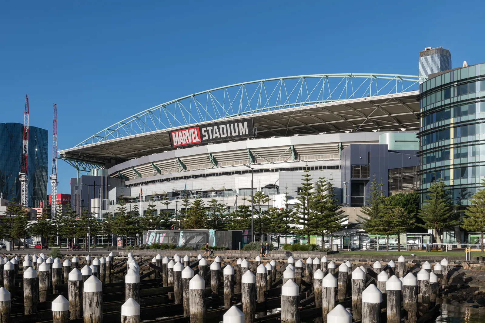 Marvel Stadium in Melbourne, viewed from across the water with its retractable roof open under a clear blue sky.