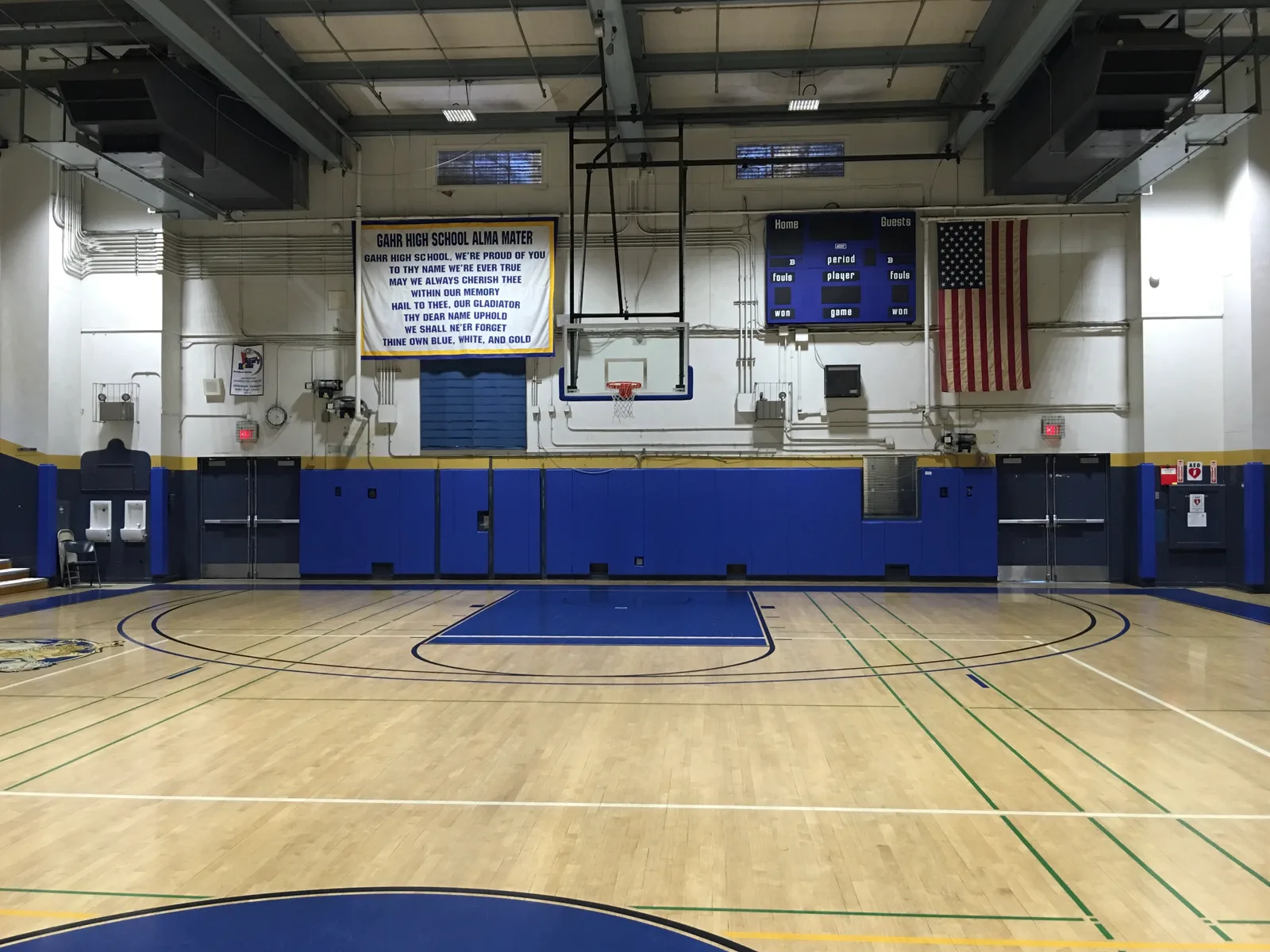An empty indoor basketball court viewed from courtside, with a hoop, scoreboard and American flag mounted on the wall.