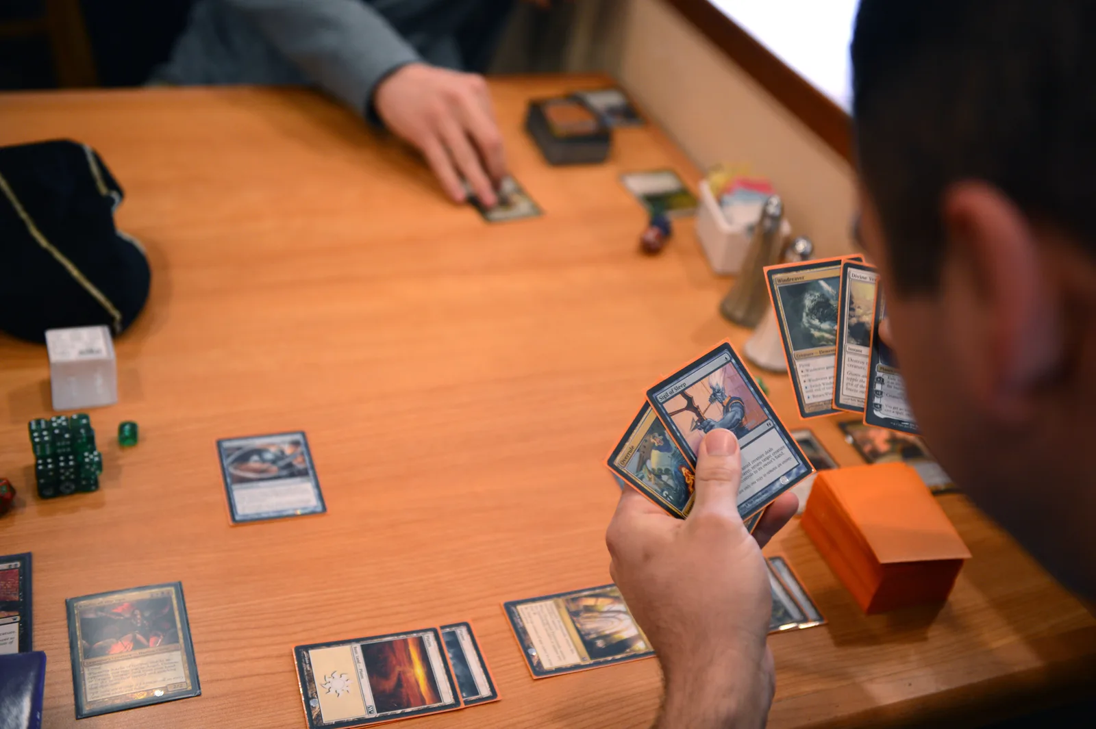 Two players examine Magic: The Gathering cards fanned out across a wooden table during a game.