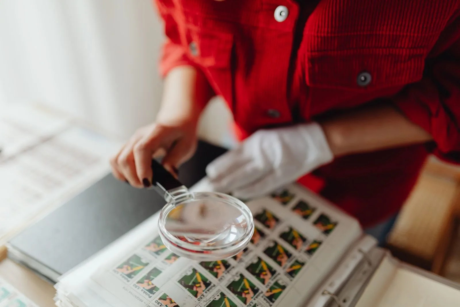 A person wearing a white cotton glove holds a magnifying glass over a page of postage stamps in a collector's album.