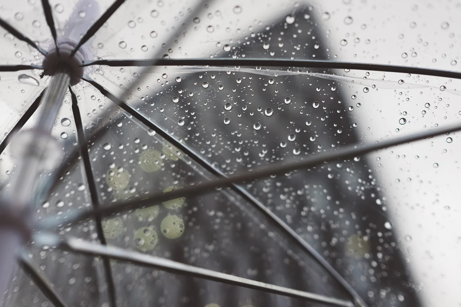 Raindrops beaded on a clear umbrella, seen from underneath against a grey sky and a blurred building.
