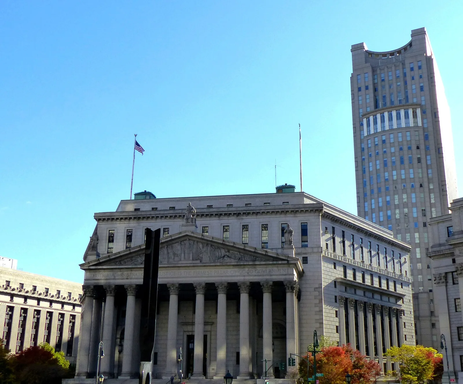 The columned facade of the New York County Courthouse in Foley Square under a clear blue sky, with the tower of the Daniel Patrick Moynihan U.S. Courthouse behind it.
