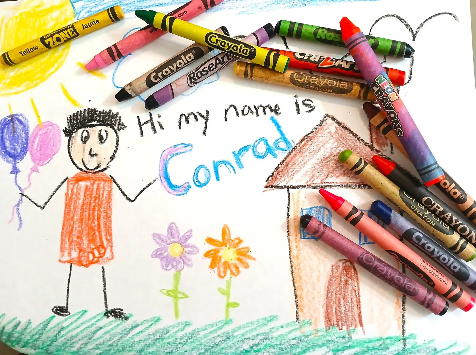 A pile of wax crayons lying on a child's crayon drawing of a smiling stick figure holding balloons, flowers and a house.