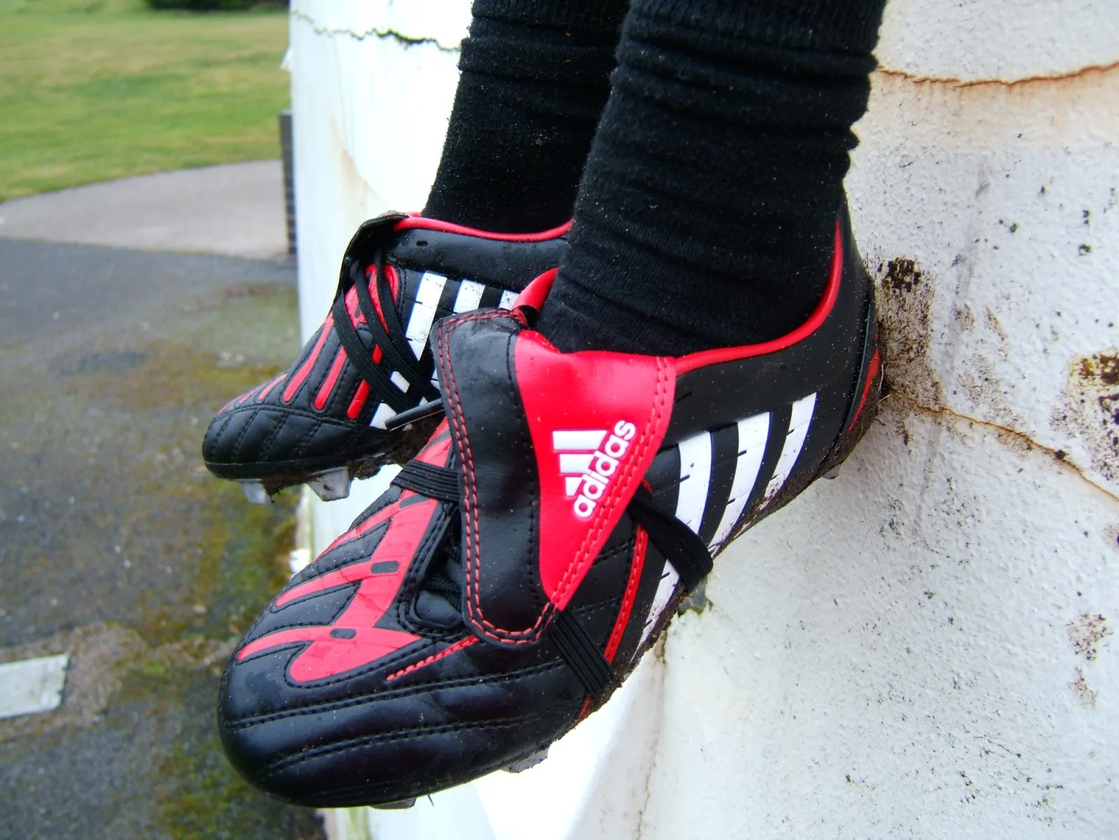 A pair of muddy black and red football boots on a player in black socks, resting against a white wall beside a grass field.