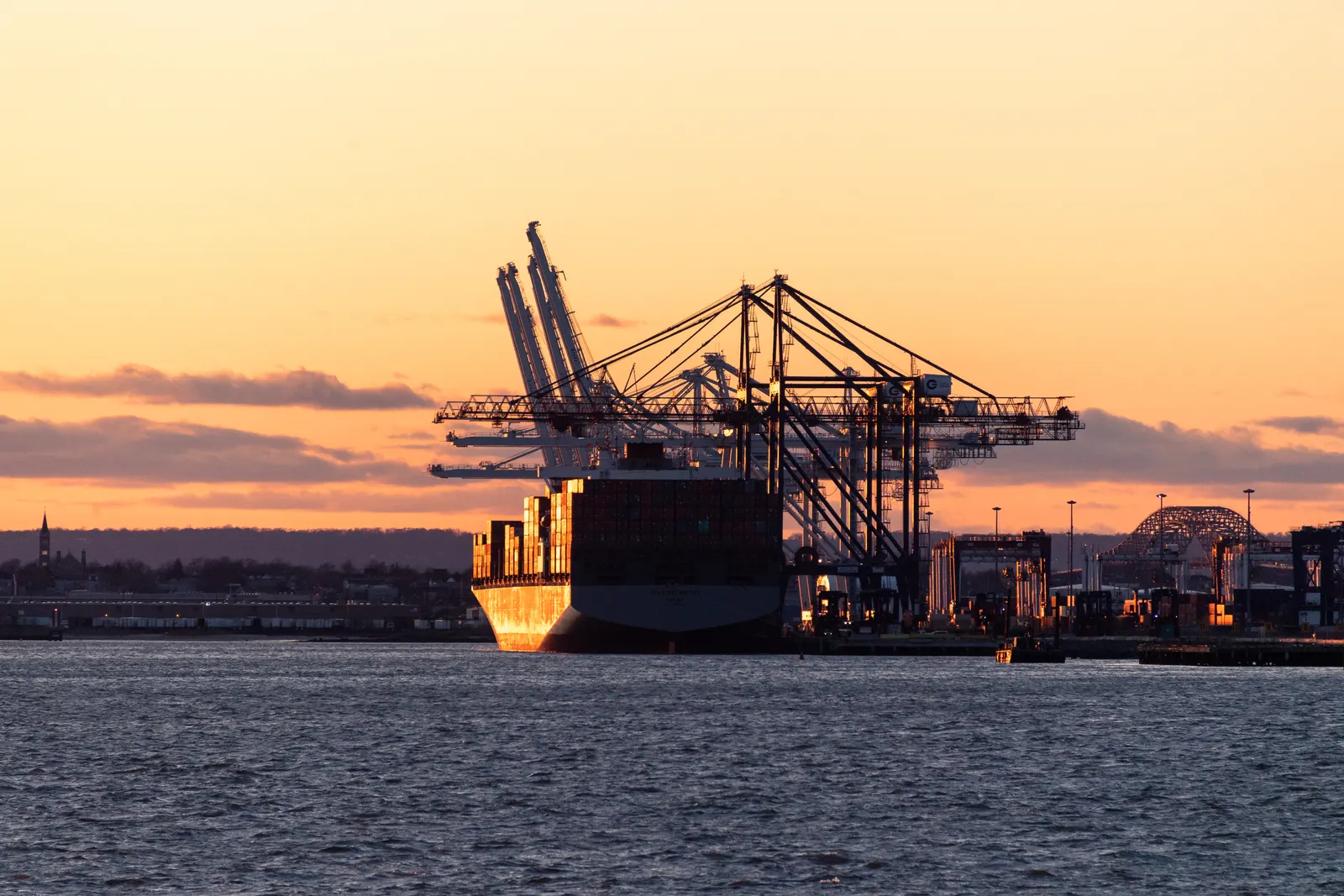 A container ship docked beside tall cranes at a port, lit by an orange sunset over the water.