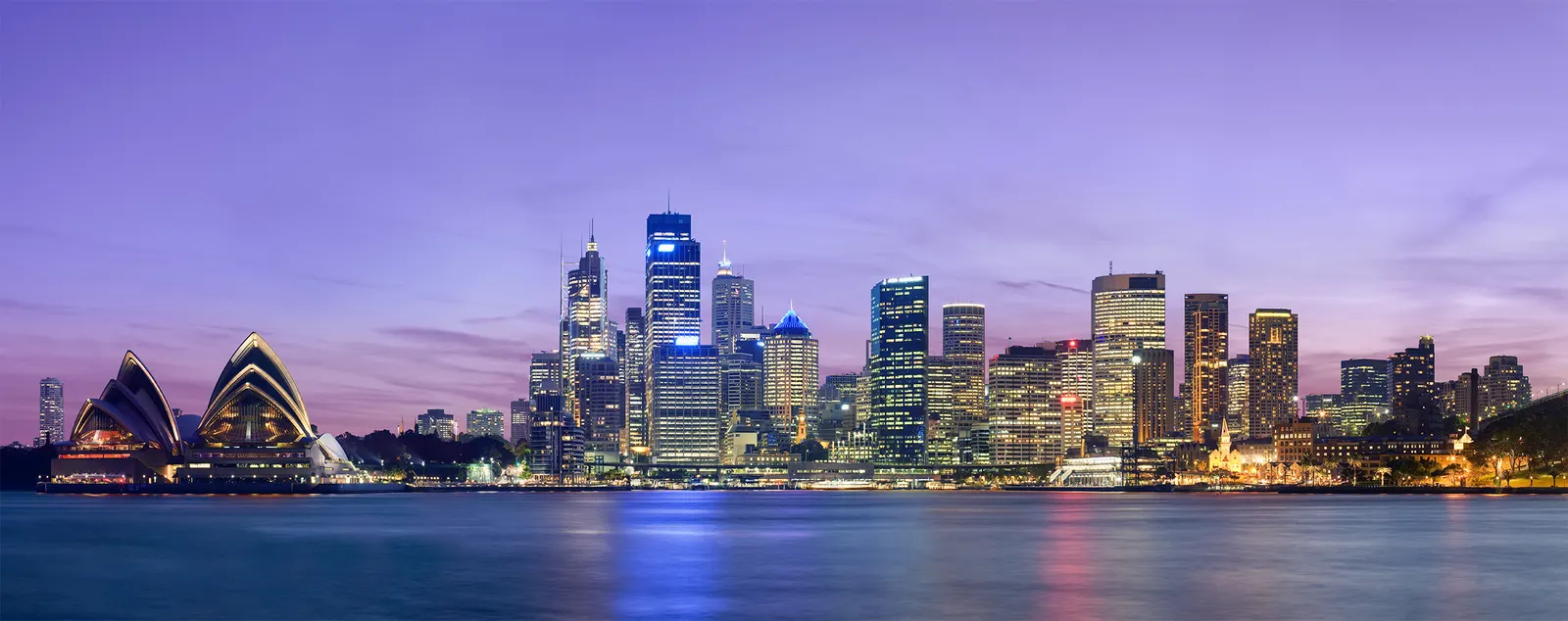 The Sydney skyline and Opera House lit up at dusk across the harbour under a purple sky.