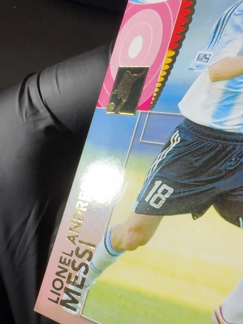 The same Lionel Messi Panini 2006 card front after the indent repair, held in a gloved hand under angled raking light at CardRevive